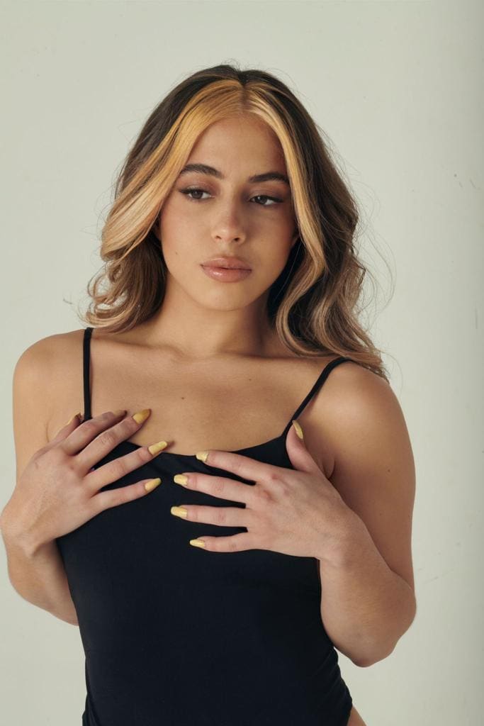 Milka with wavy brown and blonde hair wears a black spaghetti-strap top, resting hands on chest against a light background.