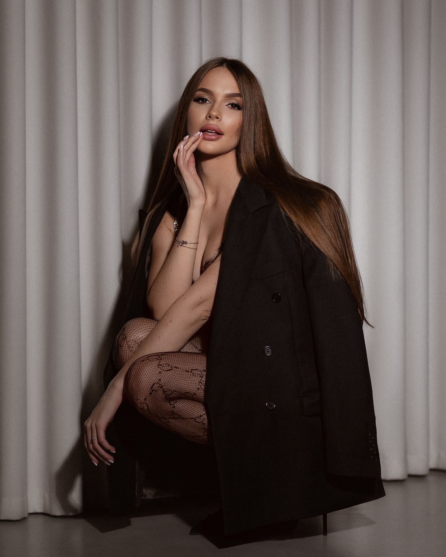Crouching before a white curtain, she wears an open black coat, dark lace bodysuit, and patterned fishnets, hand raised to her chin.