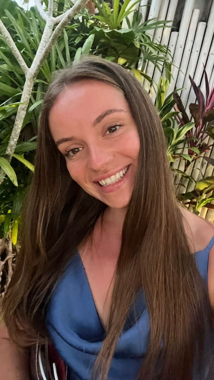 A smiling woman in a blue sleeveless top stands before green foliage and a bamboo fence.