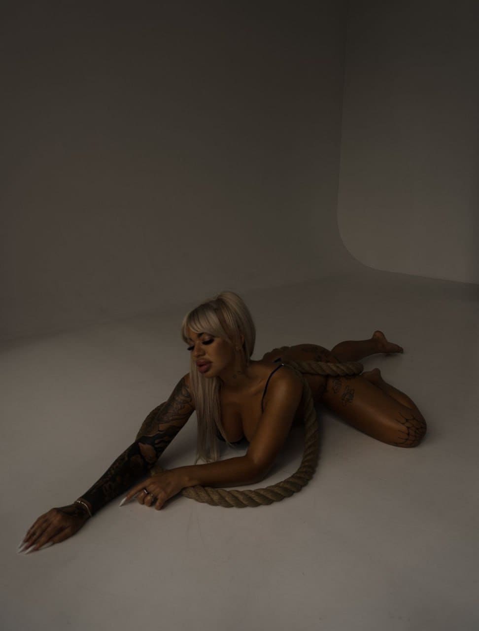 A tattooed blonde woman lies on a gray studio floor in a black bra with a rope wrapped around her torso, arm extended forward.