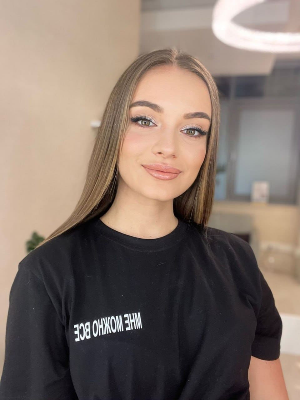 Bonnie stands indoors in a black t-shirt with white text, facing a blurred background with a ring light above.