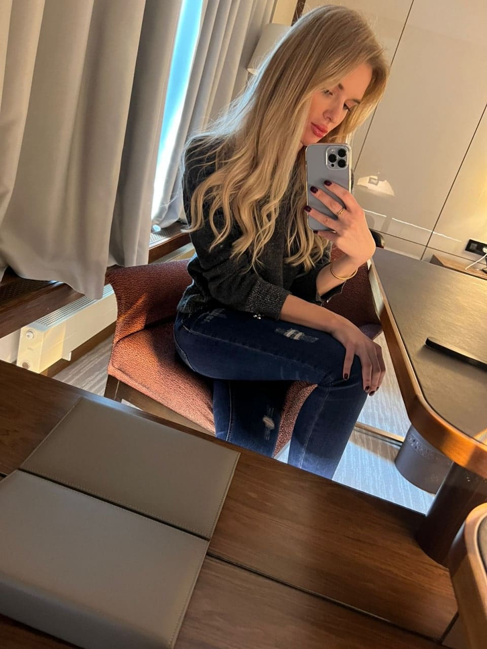 A woman with long wavy blonde hair sits on a cushioned chair, holding a smartphone over her face, wearing a dark long-sleeved top and blue jeans.