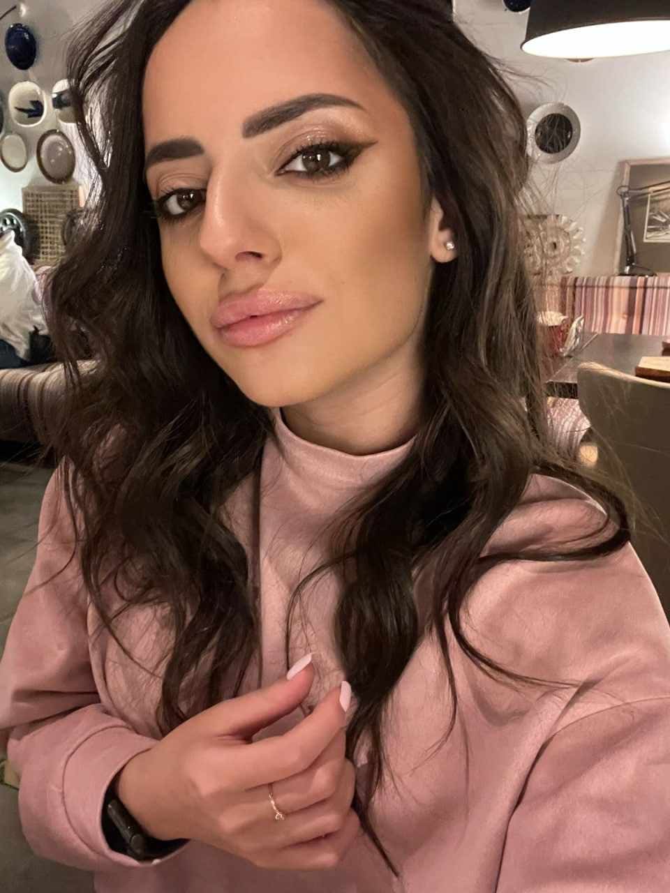 Zarina wears a pink turtleneck and winged eyeliner, posing in a close-up selfie with a hand raised near her chest against a wall with decorative plates.