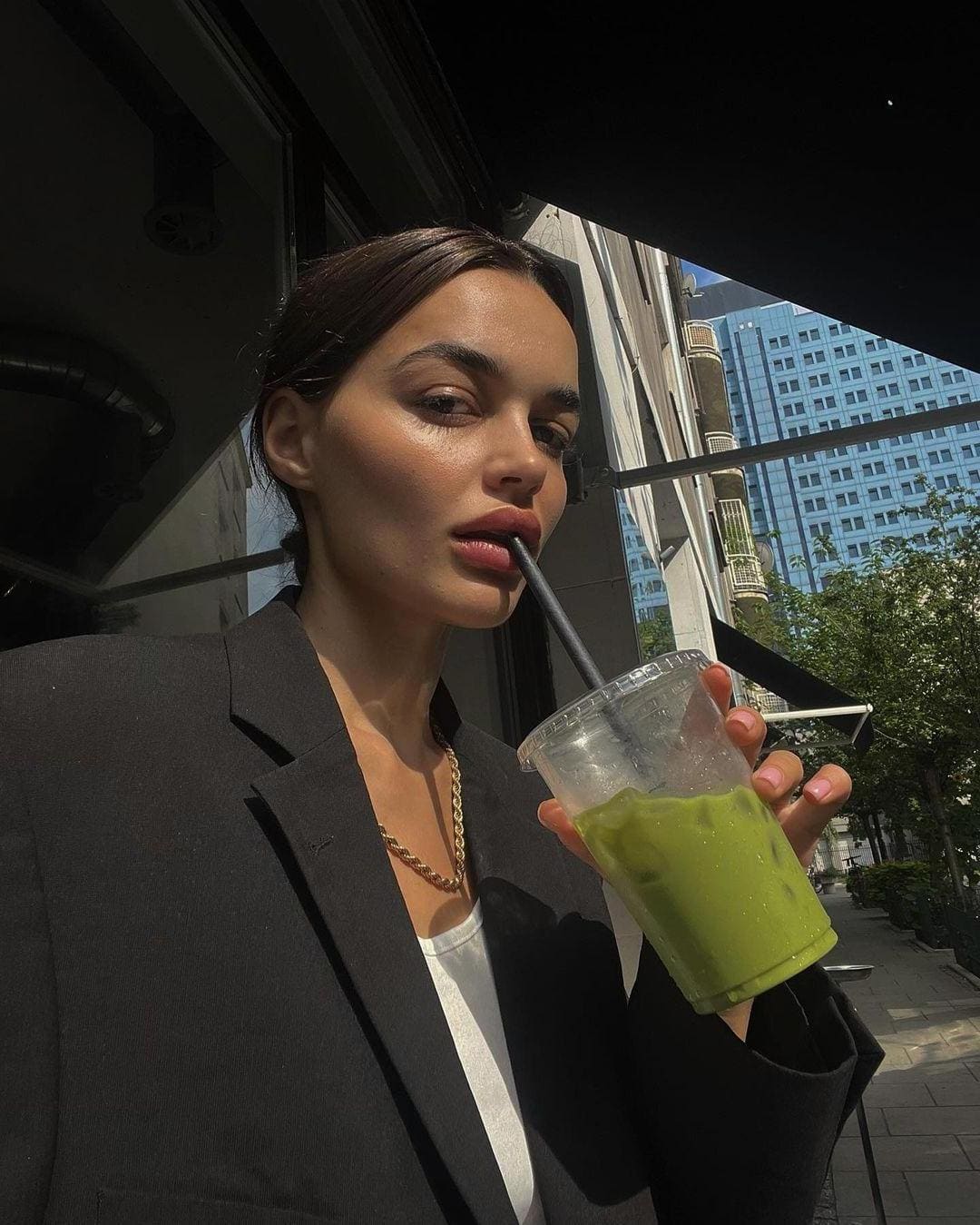 A woman in a black blazer and white top stands on a city sidewalk, holding a clear cup with a green iced drink and black straw.