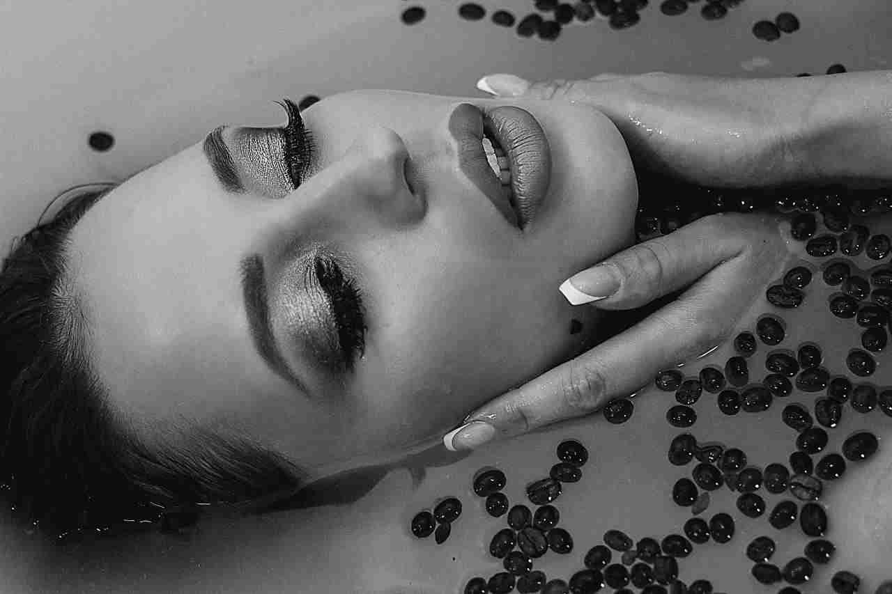 Black-and-white close-up of a woman lying in water with dark beans, eyes closed and face partially submerged, showing long nails.