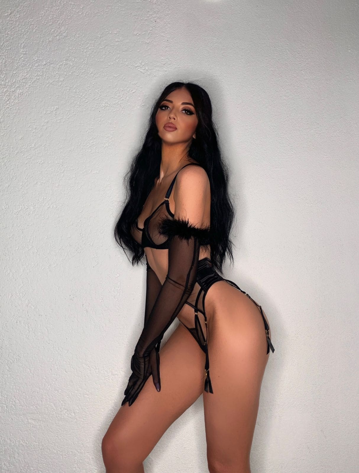 Side pose of a woman in black lace bra, sheer gloves, and garter bottoms against a white textured wall.