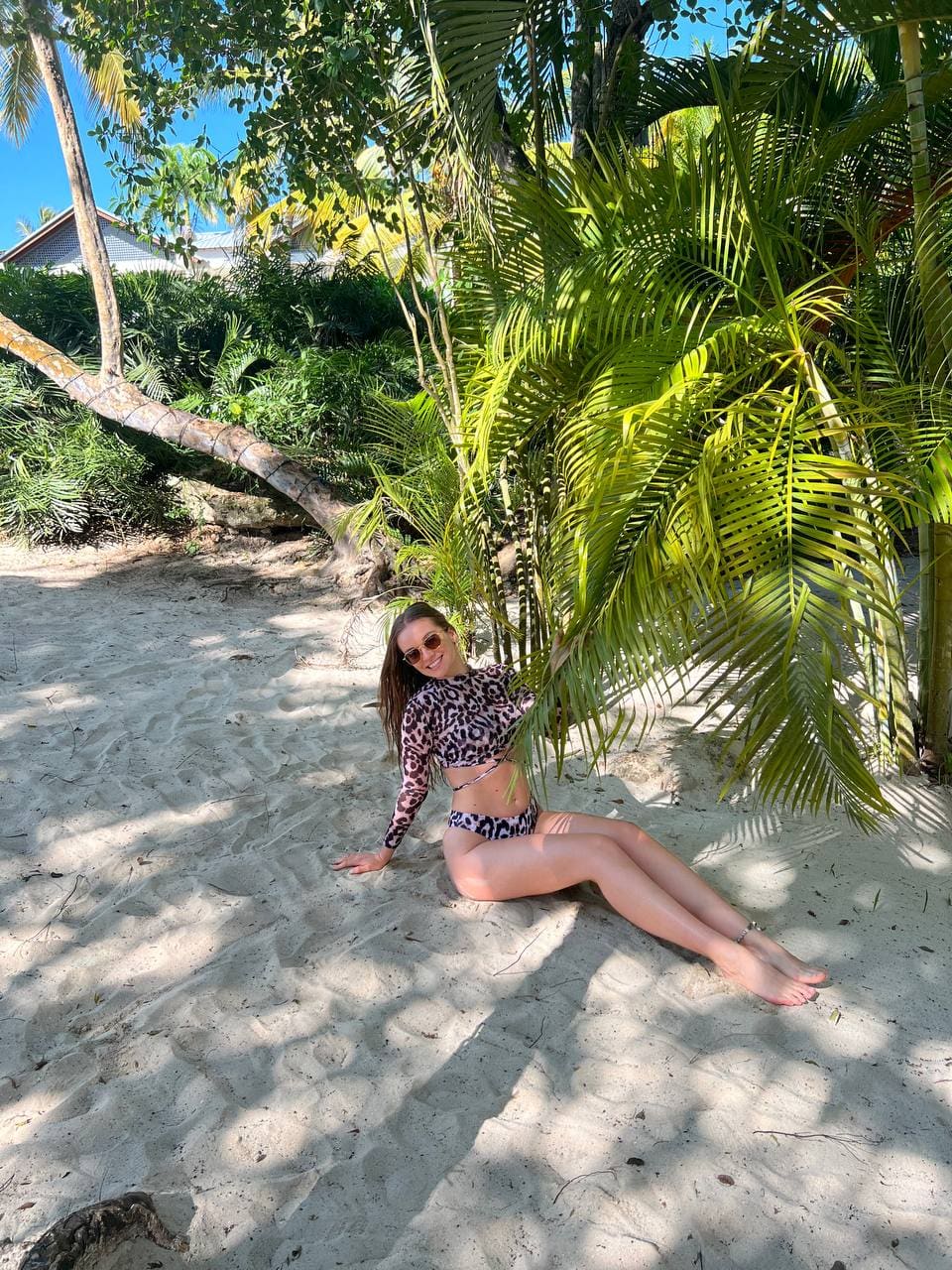 Wearing sunglasses and a leopard-print swimsuit, a woman leans against a palm tree on a sandy beach.