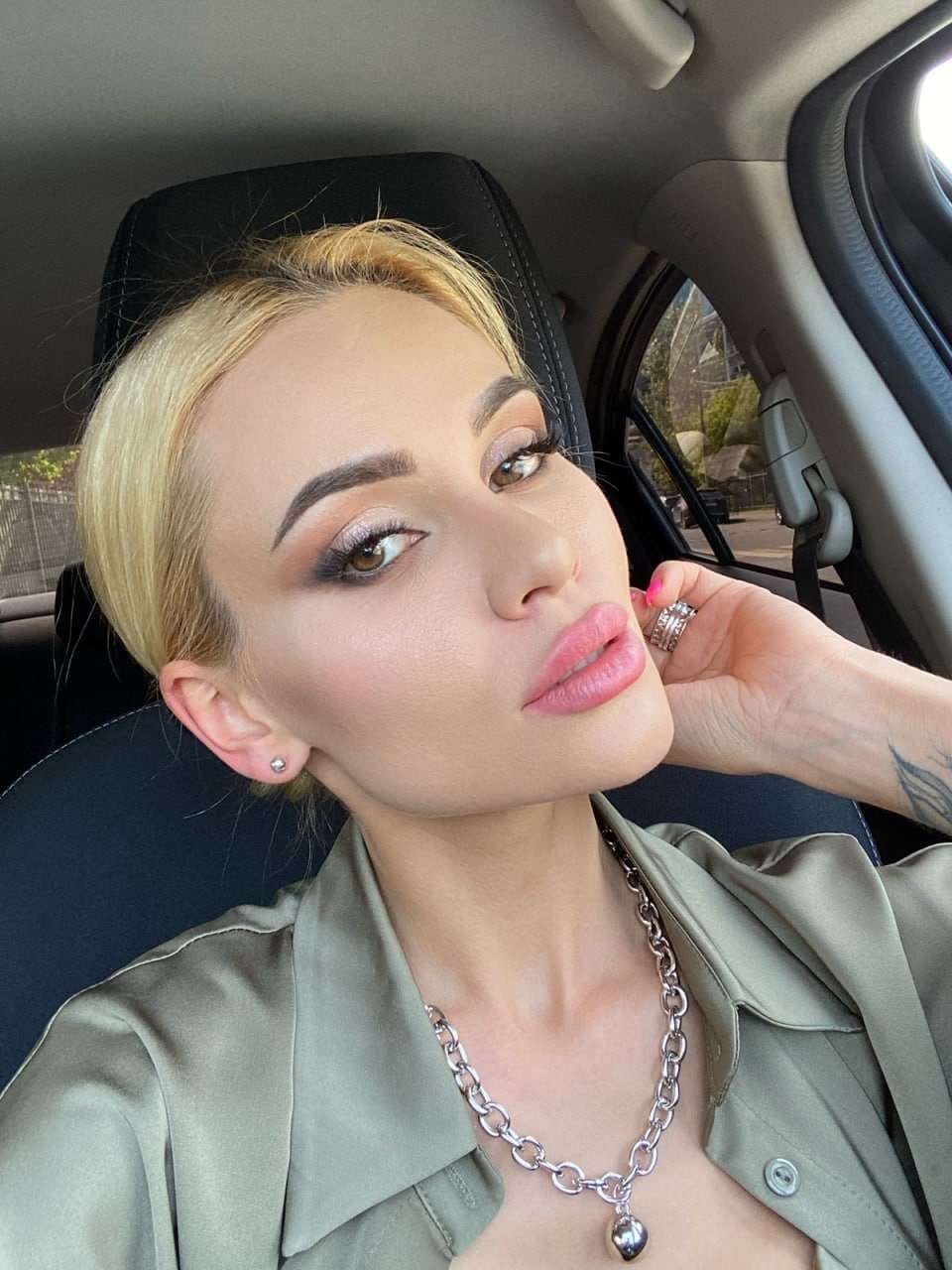 Anita sits in a car driver's seat, wearing an open olive-green shirt and silver necklace, with her hand resting near her chin.