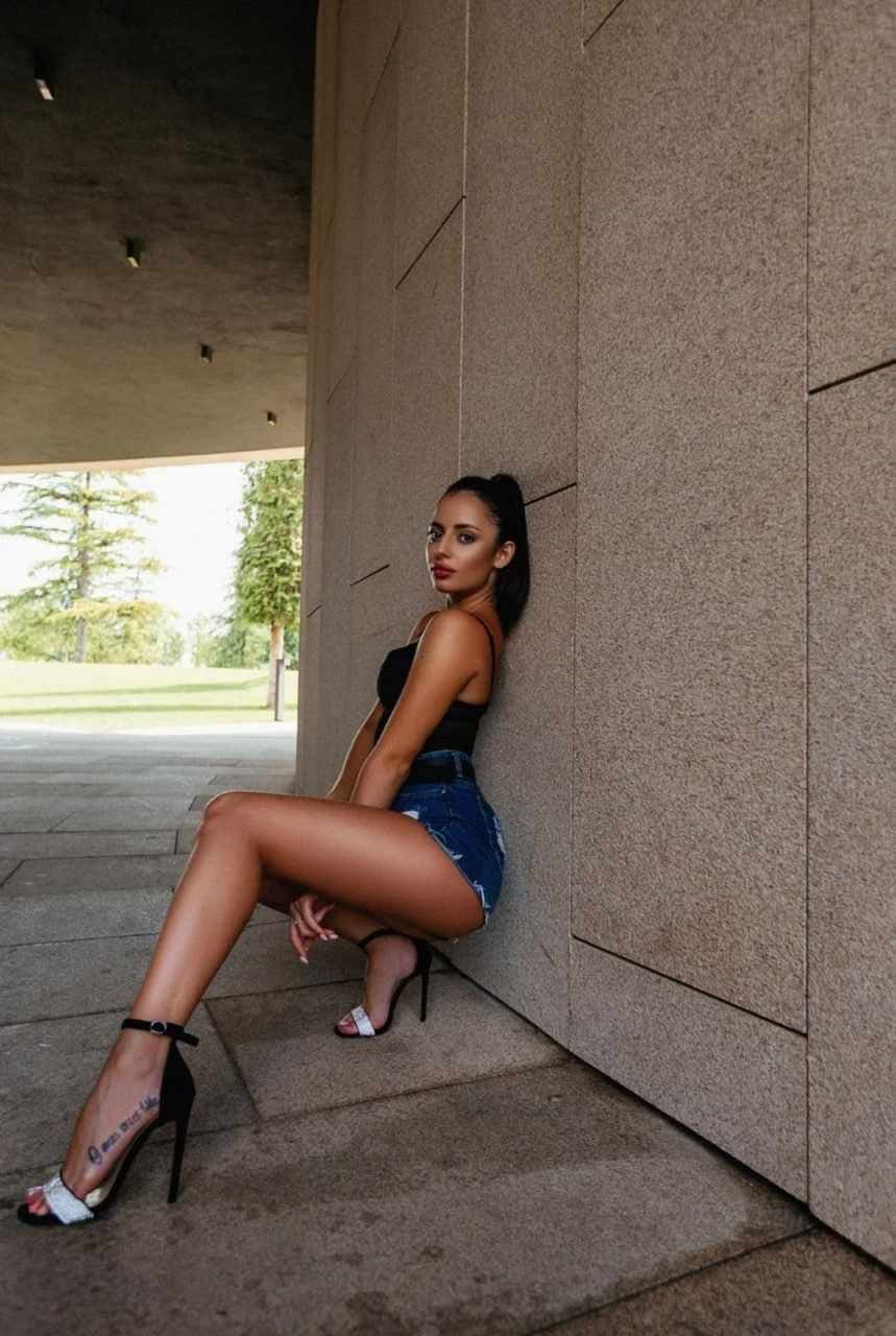A woman with a high ponytail sits on a stone ledge in a black strapless top and denim shorts, legs crossed, against a concrete wall.