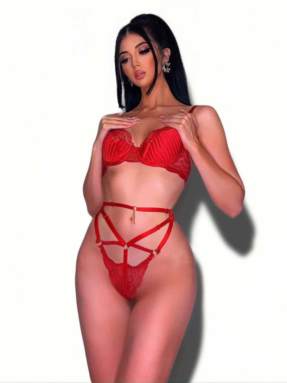 Woman in red lace bra and thong poses with hands near her chest against a plain white background.