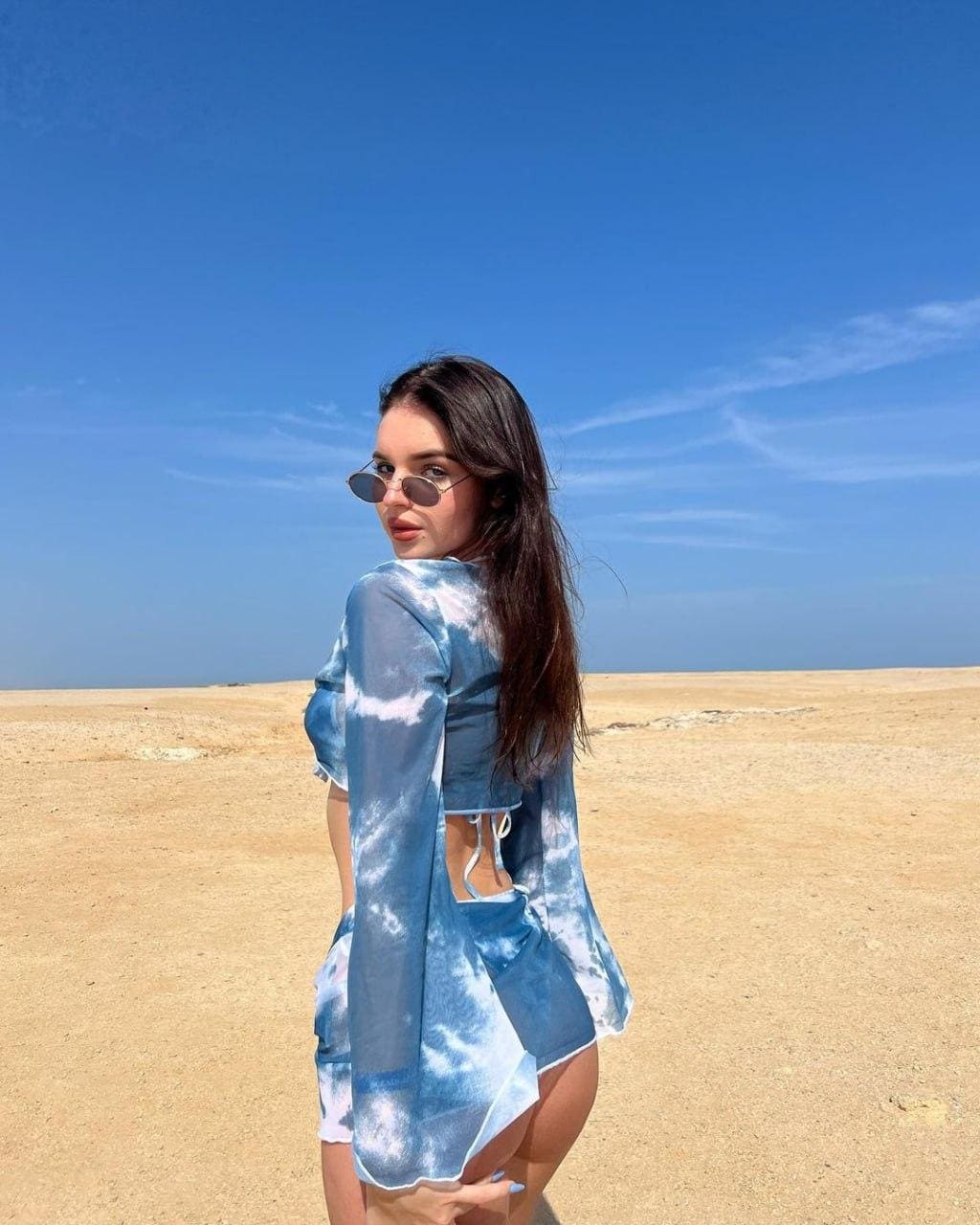 A woman in blue and white tie-dye clothing stands on a sandy beach under a clear sky, wearing round sunglasses with her back partly turned.