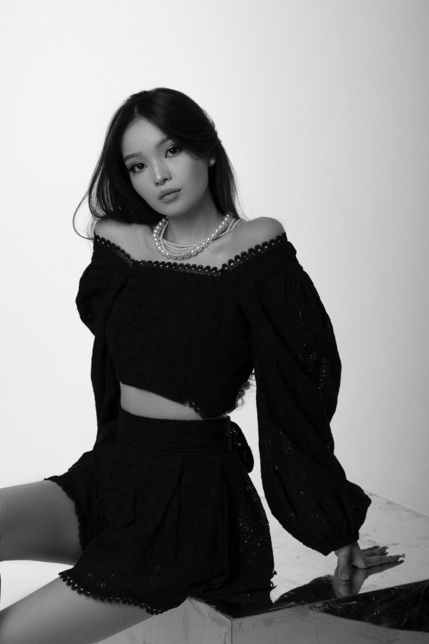 Aurika sits on a reflective surface in a black off-the-shoulder crop top and shorts with a pearl necklace.