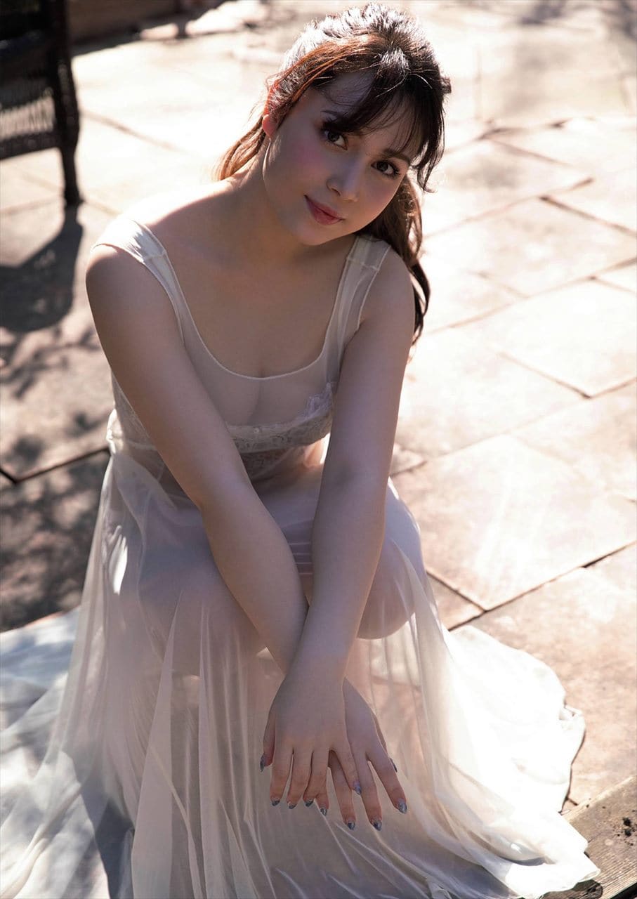 Seated on a stone patio, she wears a sheer sleeveless white dress over lace-trimmed undergarments, hands resting on her knees.