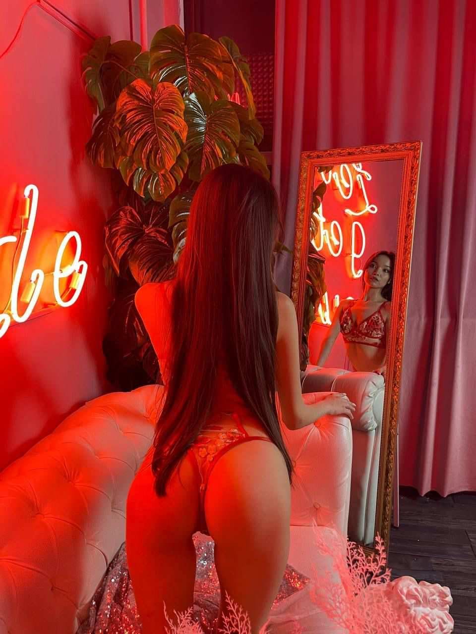 A woman in red lace lingerie kneels on a white sofa, seen from behind in red neon light.