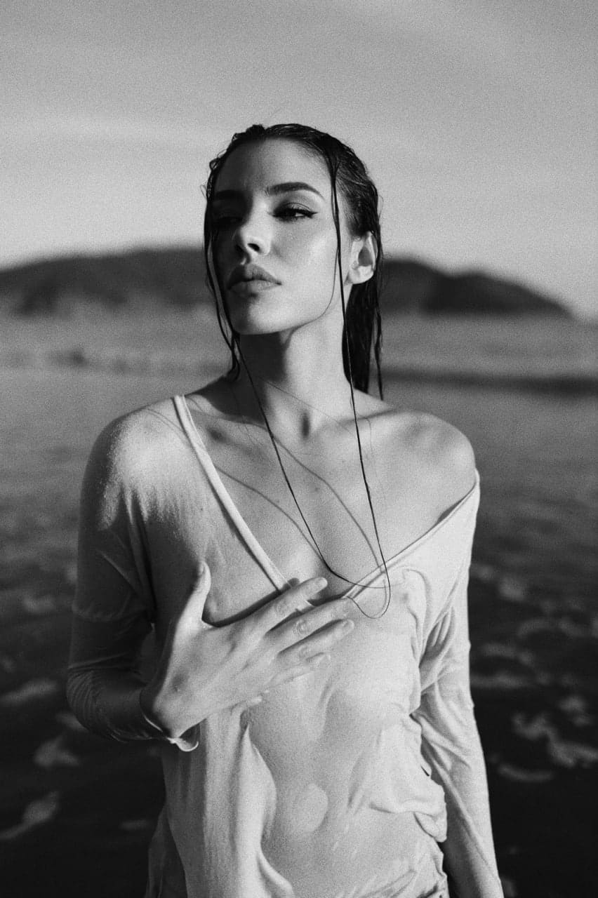 A woman with slicked-back wet hair stands near water in a clinging off-shoulder top, one hand resting on her chest.