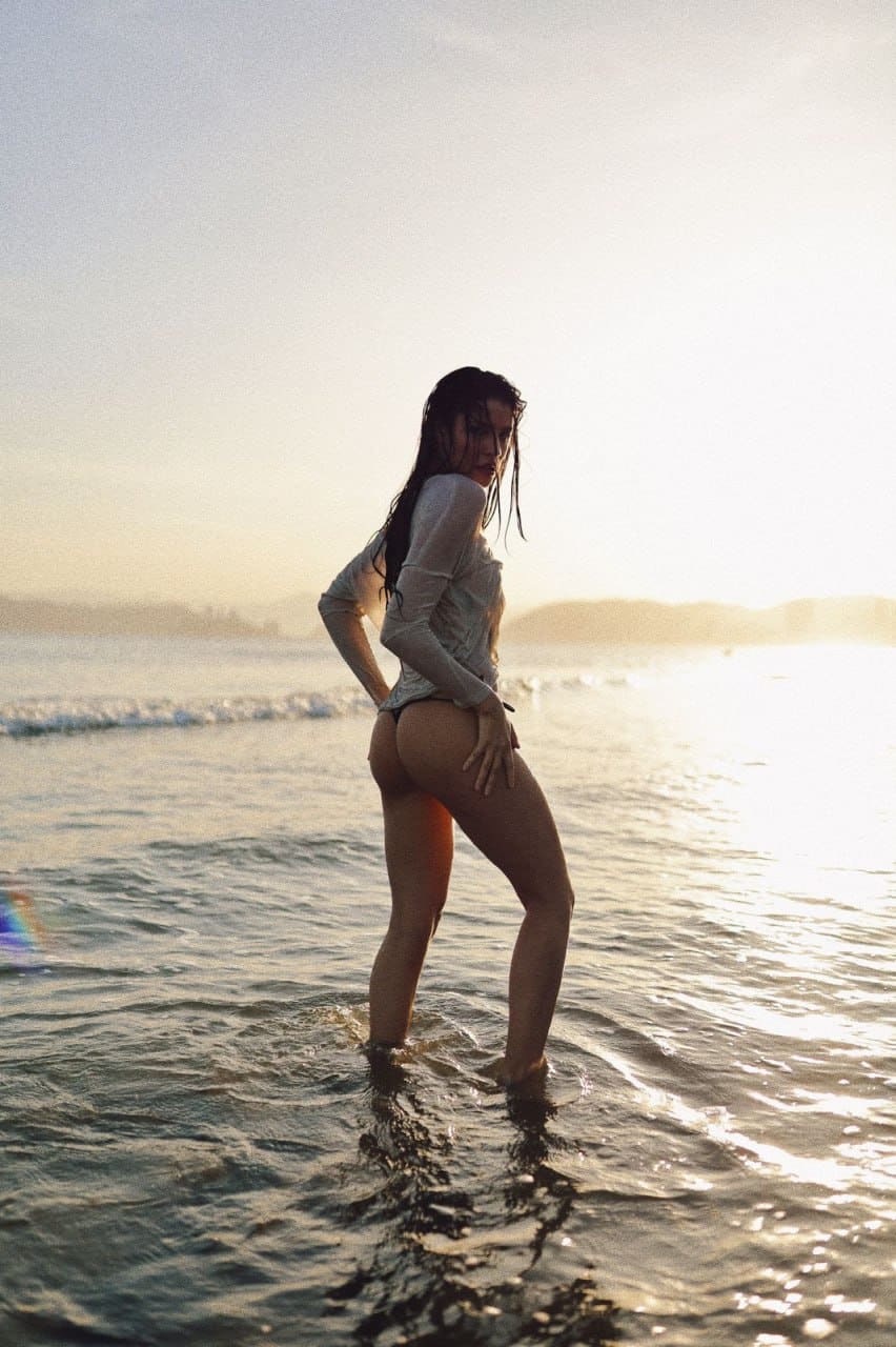 At sunset, a woman in a light long-sleeve top and bikini bottom stands in ocean water, hand on hip.