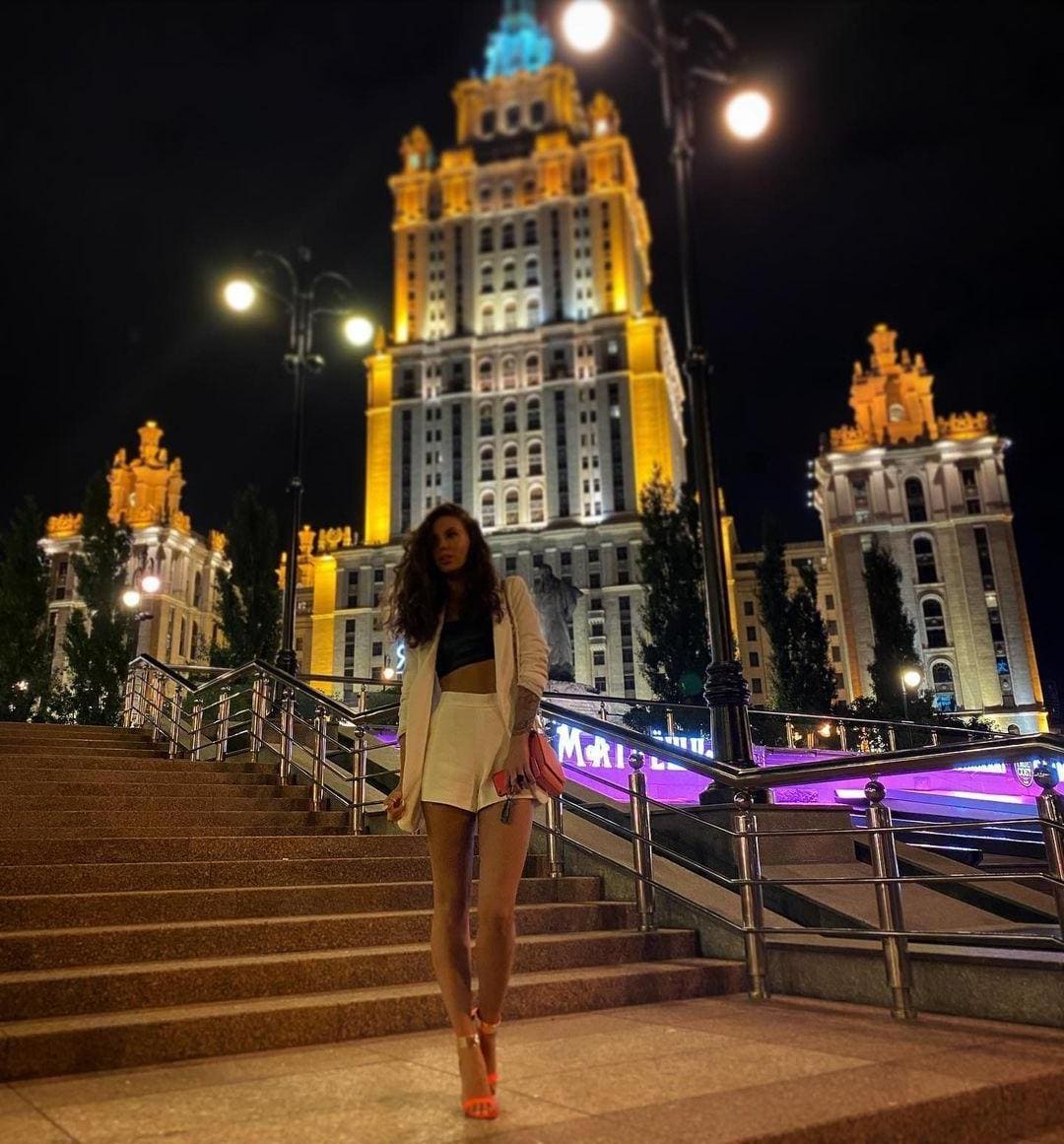 A woman in a white blazer and shorts stands on stone steps at night before an illuminated building.