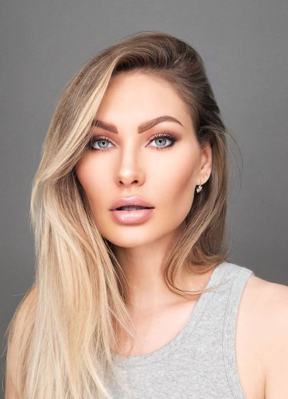 Aksena faces the camera in a grey sleeveless tank top and stud earrings, with long blonde hair against a plain grey background.