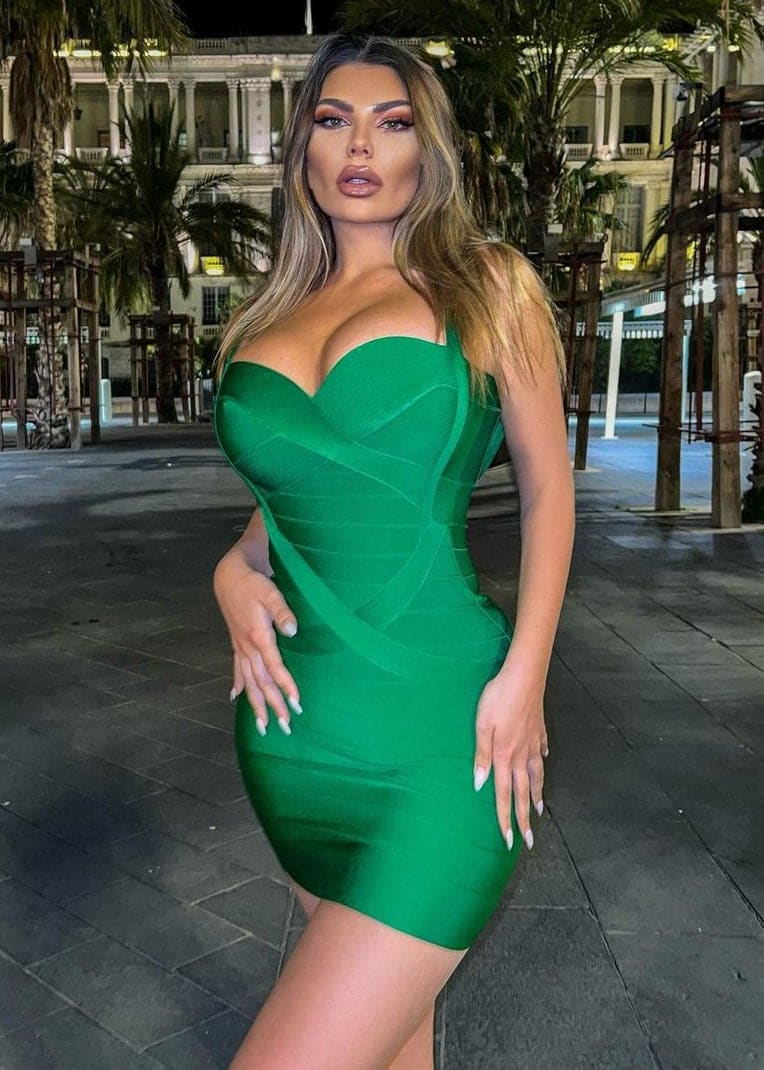 A woman in a strapless green mini dress stands at night in front of palm trees and a classical building.