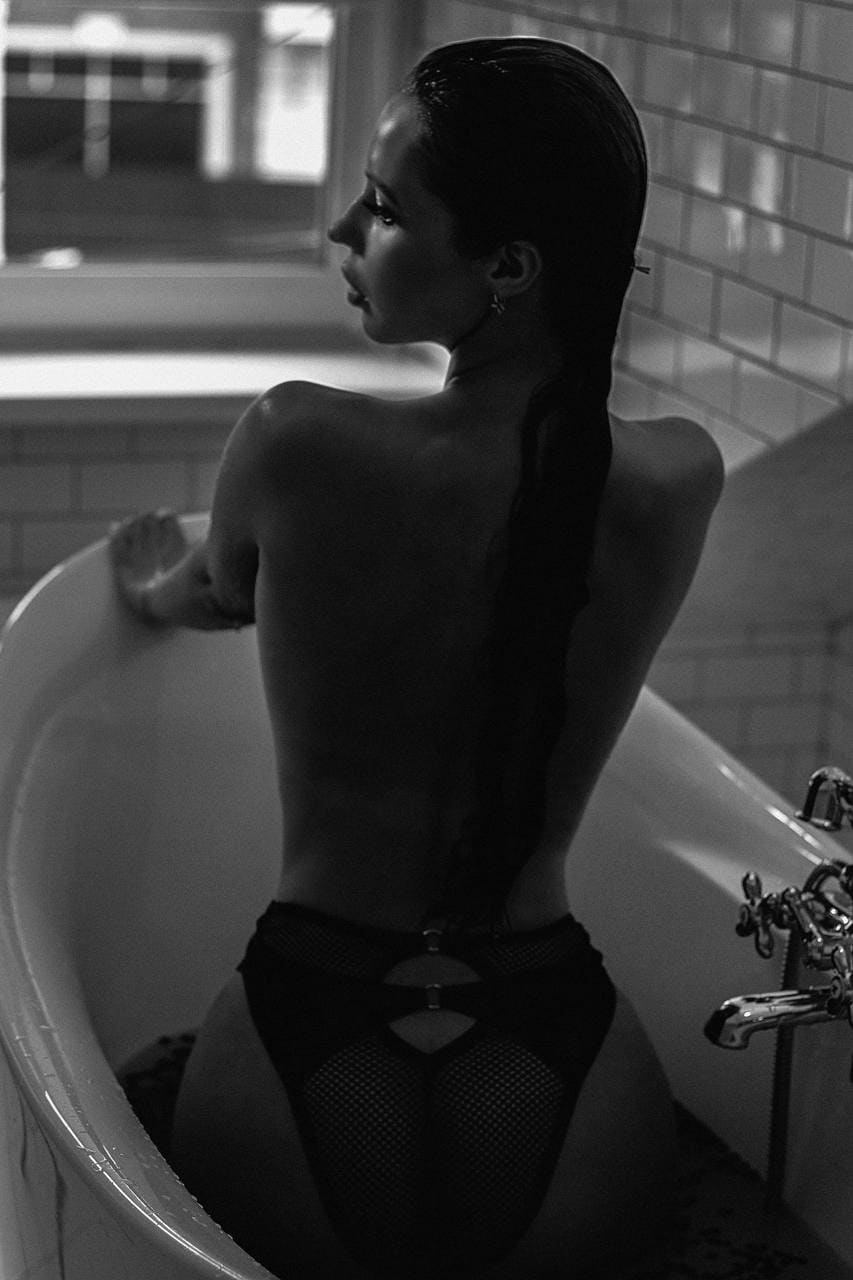 A woman with a braided hairstyle sits in a bathtub wearing a black mesh bodysuit, her bare back visible near a window.