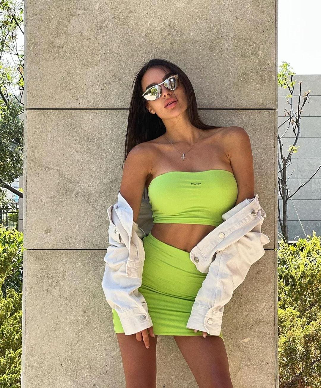 A woman in a lime green crop top and skirt leans against a pillar, wearing mirrored sunglasses and a denim jacket.