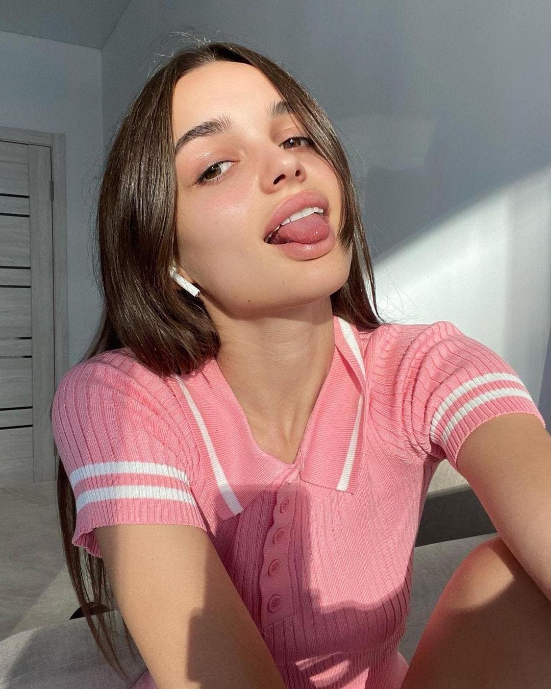 A young woman with shoulder-length brown hair takes an indoor selfie in a pink ribbed polo shirt, sticking her tongue out at the camera.