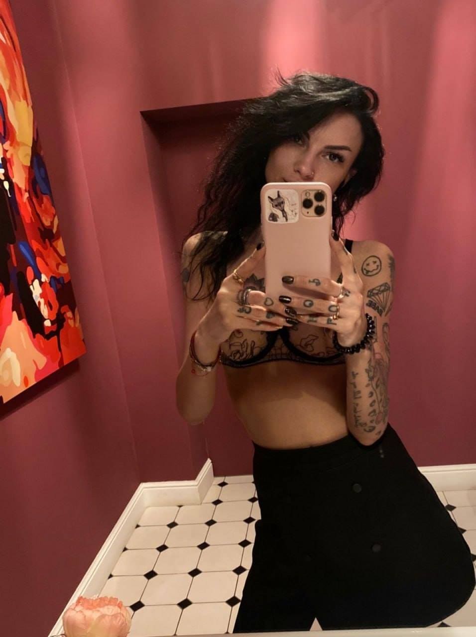 A woman with tattoos takes a mirror selfie in a pink room, wearing a black lace bra and high-waisted black skirt.