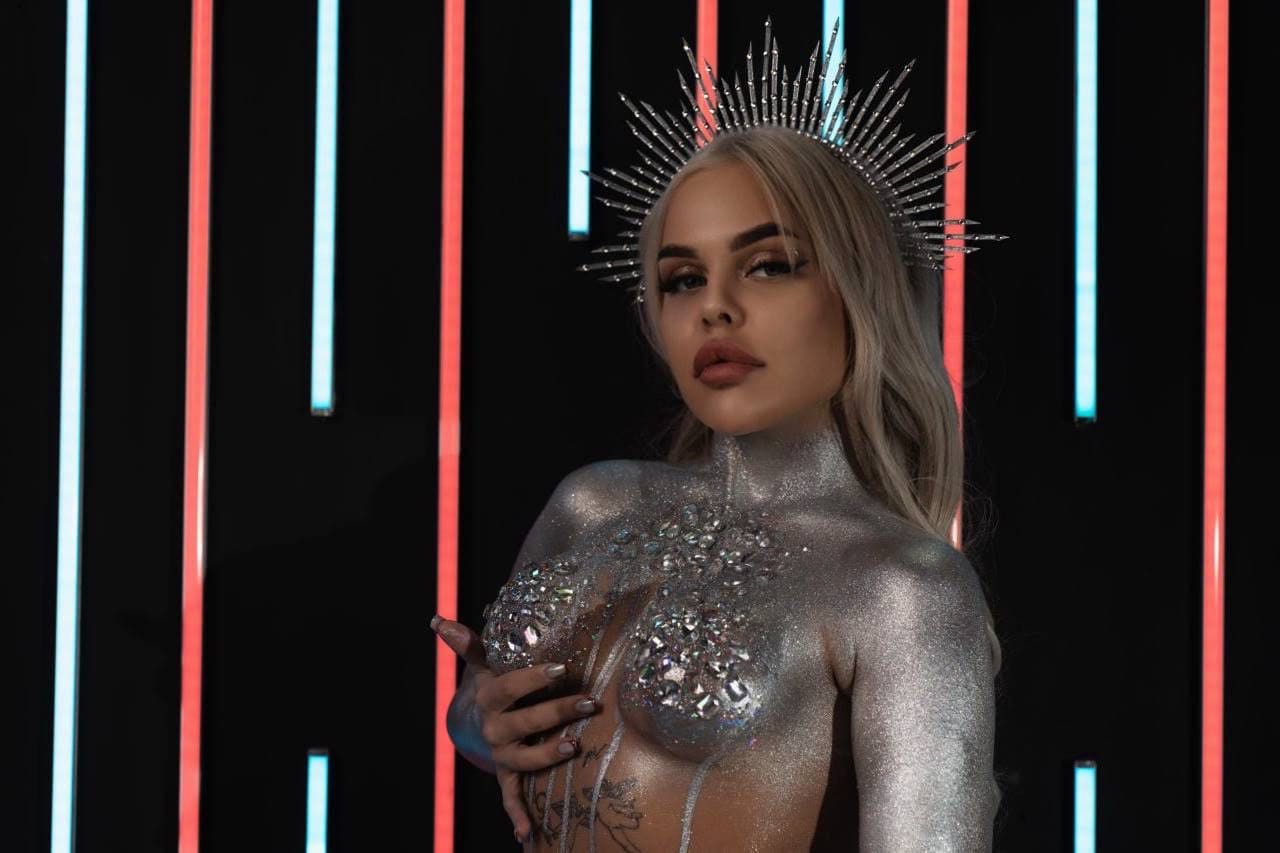 A blonde woman in a silver glitter bodysuit, jeweled bustier, and spiked crown stands before red and blue neon lights with her hand on her chest.