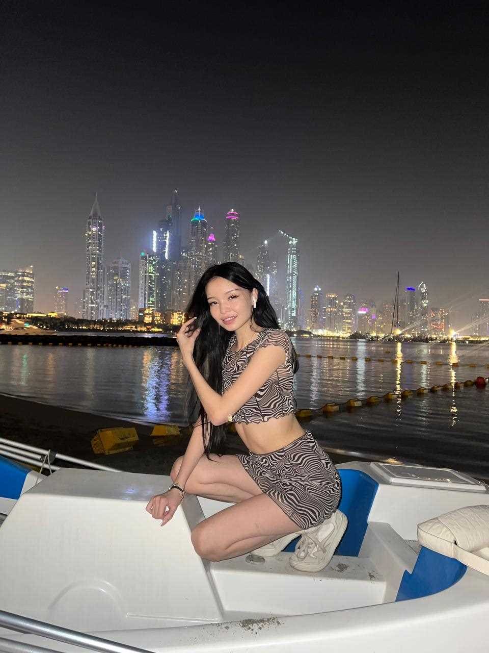 A woman in a zebra-print crop top and skirt crouches on a boat deck at night with city lights.