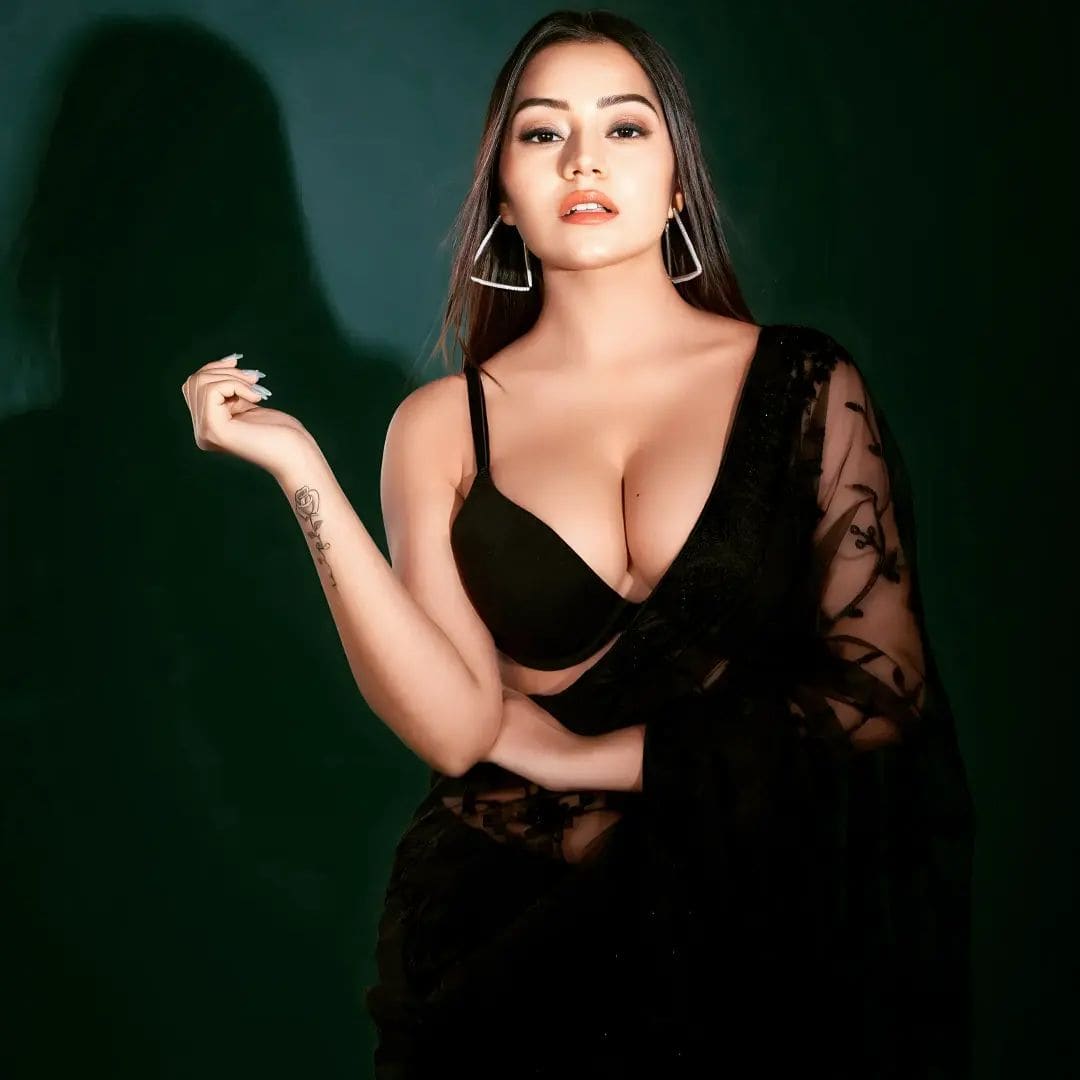 Woman stands against a dark green backdrop in a black bra and sheer lace wrap, one arm raised revealing a small forearm tattoo.