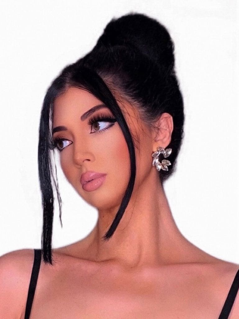 Close-up of a woman with a high bun and face-framing strands wearing a black top and large crystal earring on white.