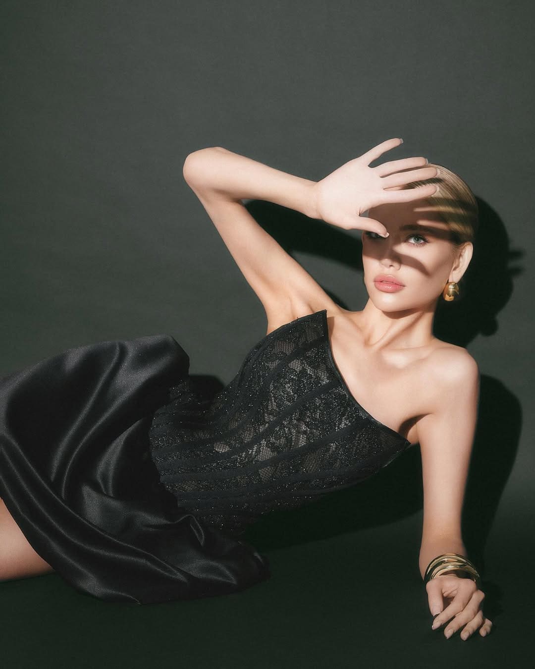 A blonde woman lies on a dark surface in a one-shoulder black lace dress, one hand raised over her forehead against a dark background.