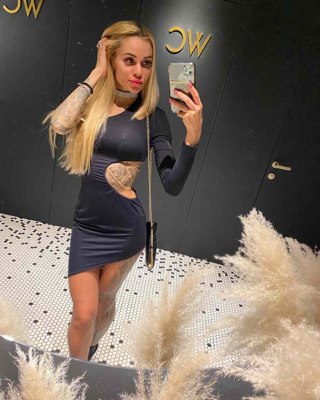 A blonde woman takes a mirror selfie in a dark mini dress with a side cutout, standing on a black-and-white tiled floor.