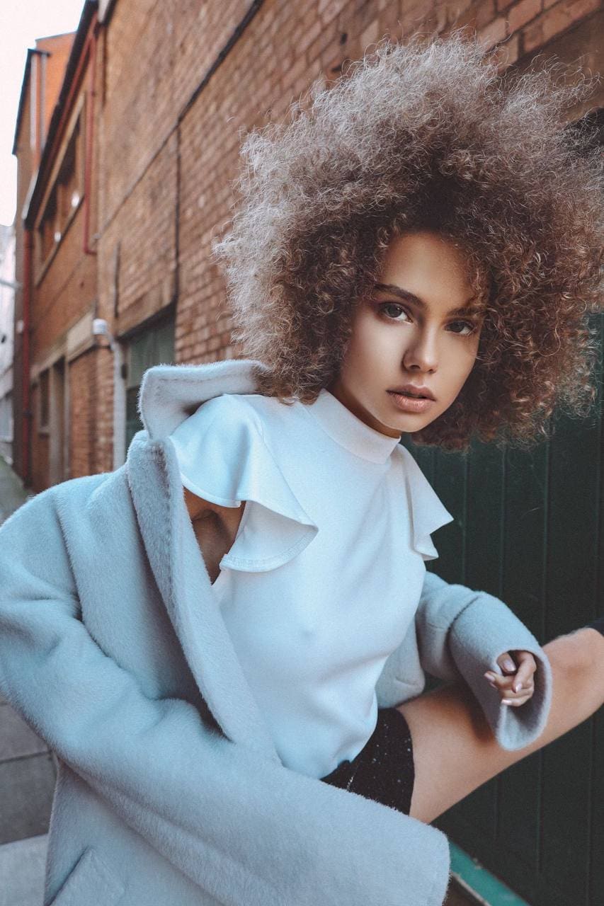 Outdoors against a brick wall, she wears a white ruffled top and light blue coat off-shoulder, one leg raised and bent.